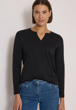 Femme aux cheveux courts et bruns portant un haut noir à manches longues avec un décolleté en V et un jean bleu sur un fond clair uni.