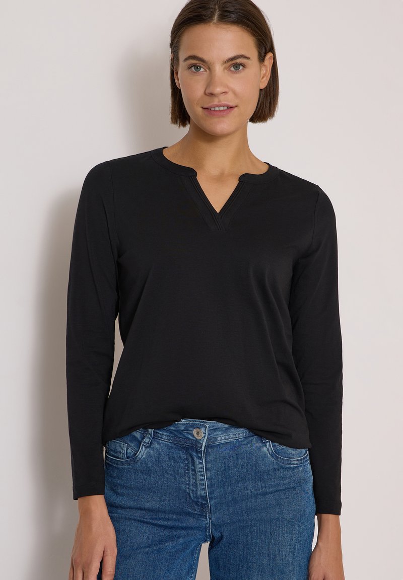 Femme aux cheveux courts et bruns portant un haut noir à manches longues avec un décolleté en V et un jean bleu sur un fond clair uni.
