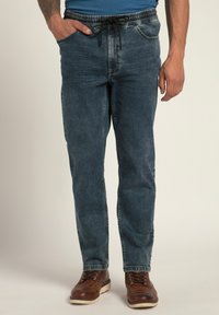 Vaqueros de mezclilla en azul oscuro con una cinturilla elástica y cordón. Presentan bolsillos laterales y un ajuste relajado. Unos zapatos de cuero marrón complementan el look.