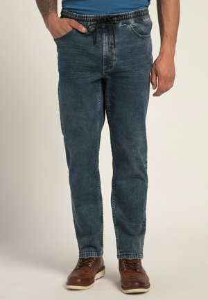 Vaqueros de mezclilla en azul oscuro con una cinturilla elástica y cordón. Presentan bolsillos laterales y un ajuste relajado. Unos zapatos de cuero marrón complementan el look.