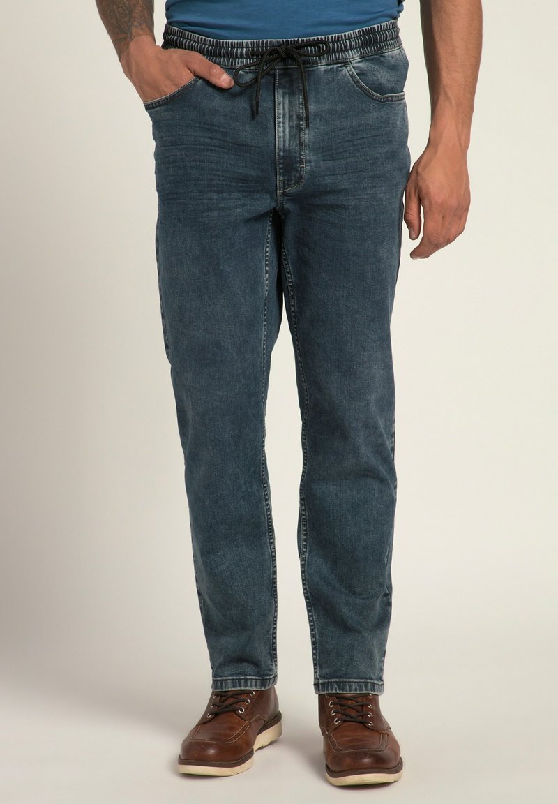 Vaqueros de mezclilla en azul oscuro con una cinturilla elástica y cordón. Presentan bolsillos laterales y un ajuste relajado. Unos zapatos de cuero marrón complementan el look.