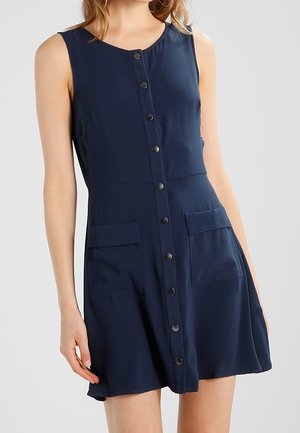 Sleeveless navy dress with front button closure and two flap pockets, worn by a woman with loose hair against a plain background.