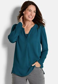 Türkise Bluse mit langen Ärmeln und V-Ausschnitt, aus glattem Stoff gefertigt. Mit seitlichen Schlitzen und lässiger Passform. Graue Hose sichtbar.