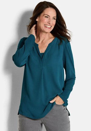 Türkise Bluse mit langen Ärmeln und V-Ausschnitt, aus glattem Stoff gefertigt. Mit seitlichen Schlitzen und lässiger Passform. Graue Hose sichtbar.
