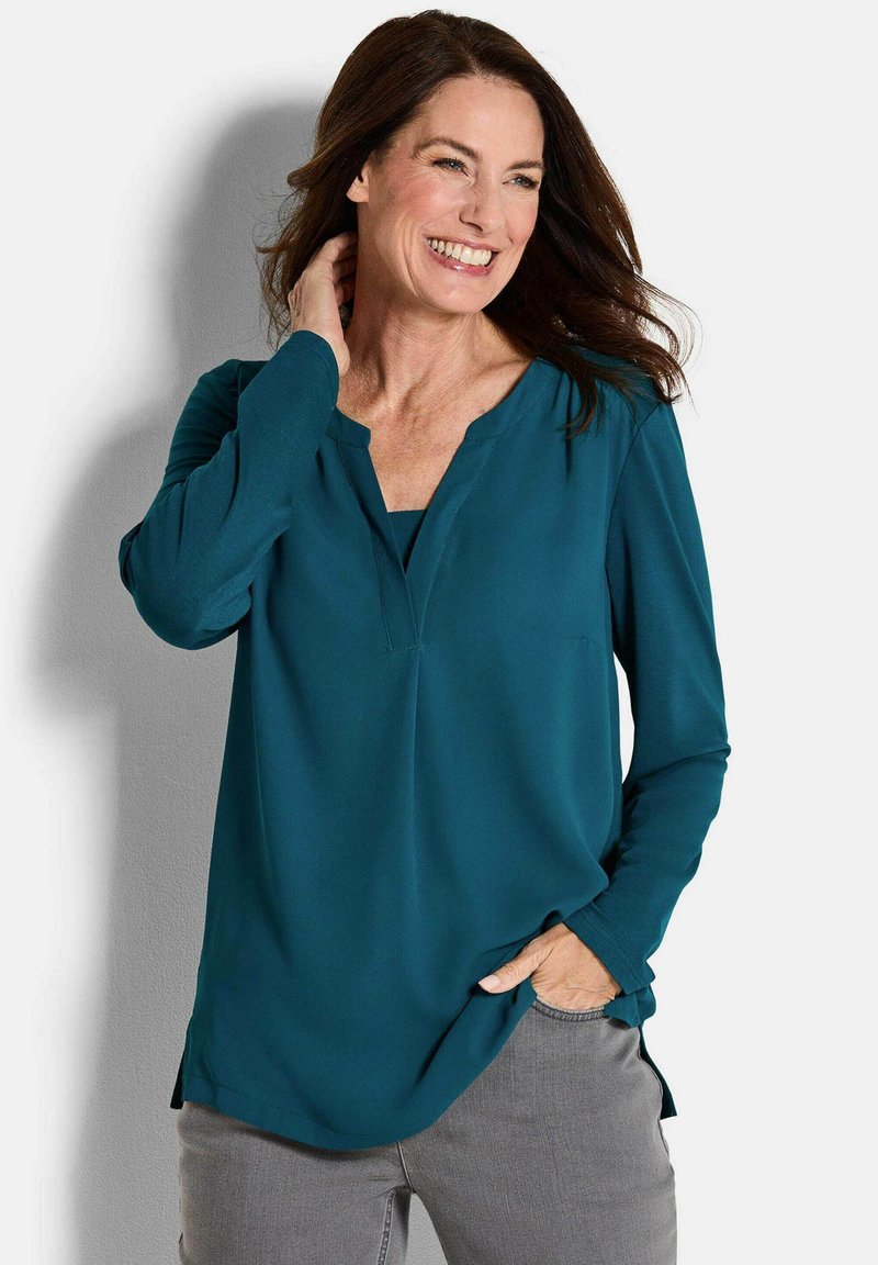 Türkise Bluse mit langen Ärmeln und V-Ausschnitt, aus glattem Stoff gefertigt. Mit seitlichen Schlitzen und lässiger Passform. Graue Hose sichtbar.