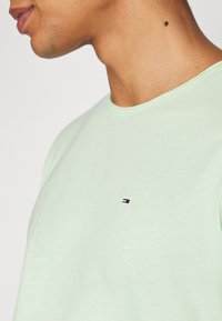 Svěže zelené tričko s krátkým rukávem a kulatým výstřihem, na hrudi s malým logem a červenými, bílými a modrými akcenty. Jemná textura.