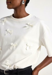 Hvid bomulds t-shirt med afslappet pasform, prydet med flere små cremede satinbånd og rund halsudskæring, parret med et sort bælte.