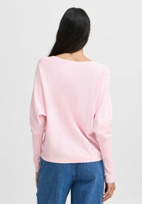 Pull léger rose pâle avec un large col, coupe décontractée et manches longues ; texture douce, poignets et ourlet côtelés. Associé à un jean bleu.