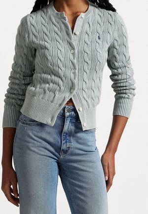 Persoon draagt een lichtgrijze kabelgebreide cardigan over een grijs shirt, gecombineerd met hooggetaillede blauwe jeans, tegen een effen achtergrond.