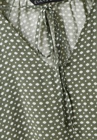 Grün gemusterte Bluse mit V-Ausschnitt, leichtem Stoff und weißen Rauten. Bänder am Hals sorgen für ein zusätzliches Detail im Design.