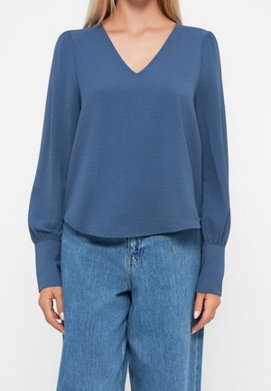 Frau trägt eine blaue langärmelige V-Ausschnitt-Bluse mit gekrempelten Ärmeln und hellblaue hoch taillierte Jeans.