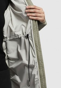 Hand som justerar den svarta dragsnöret inuti det ljusgrå foderet av en khaki jacka.