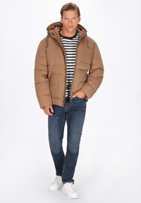 Brun oppustelig jakke med hætte, lynlås foran og lommer, båret over en sort og hvid stribet skjorte, parret med blå jeans og hvide sneakers.