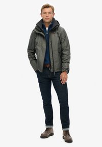 Grijze waterdichte jas met een capuchon, voorzien van ritszakken, over een blauwe fleece top. Gepaard met donkere jeans en bruine laarzen.