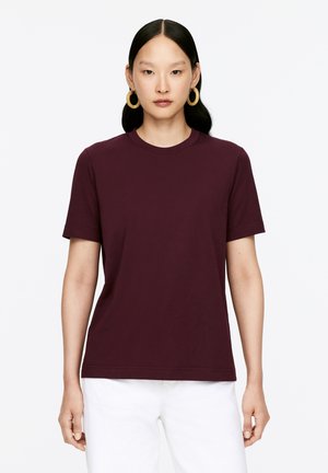 Short-sleeve, round-neck t-shirt in dark burgundy fabric. Smooth texture with a relaxed fit, paired with white pants.