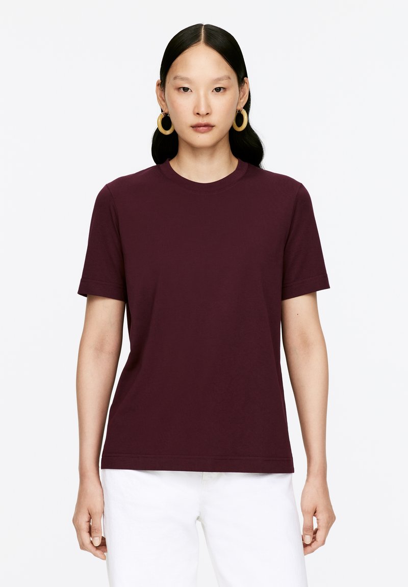 Short-sleeve, round-neck t-shirt in dark burgundy fabric. Smooth texture with a relaxed fit, paired with white pants.