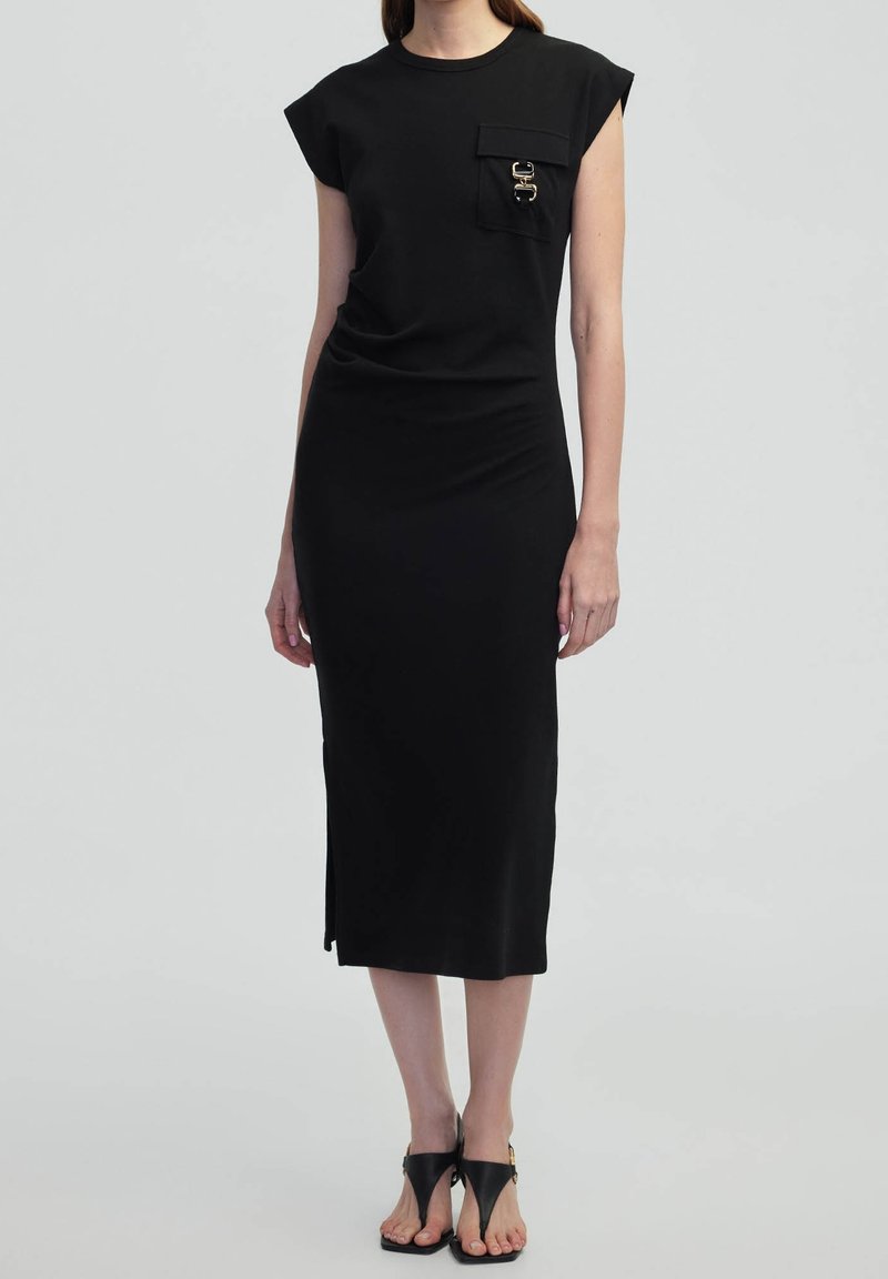Femme portant une robe midi noire sans manches avec une poche poitrine ornée de détails dorés, associée à des sandales à talons noires.