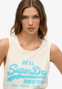 Beżowa bezrękawnikowa koszulka z niebieskim napisem "SuperDry" oraz japońskimi znakami, okrągłym dekoltem i miękką, gładką fakturą.