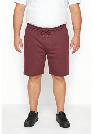 Hombre con pantalones cortos granate con cordón, camiseta blanca y zapatillas negras de pie contra un fondo blanco liso.