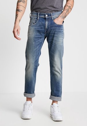 Mann mit hochgekrempelten, blau ausgewaschenen Jeans und weißen Sneakern, mit tätowierten Unterarmen und einer Hand in der Tasche vor einfarbigem Hintergrund.