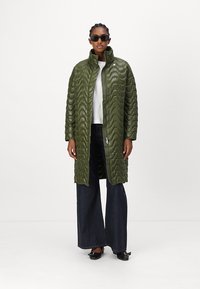 Grüne, gestepfte Puffjacke mit hohem Kragen und Frontreißverschluss, kombiniert mit einem weißen Oberteil und dunklen, weiten Jeans. Verfügt über ein strukturiertes, wellenförmiges Muster.