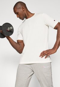 Hellbeige Baumwoll-T-Shirt mit Rundhalsausschnitt, kurzen Ärmeln und Champion-Logo am Ärmel. Graue Sporthose mit weißem Logo auf dem Oberschenkel.