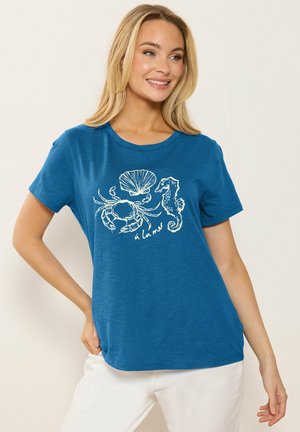Blonde vrouw draagt een blauw T-shirt met witte schetsen van zeedieren: krab, schelp, zeepaardje en de tekst "à la mer," glimlacht, hand op heup.