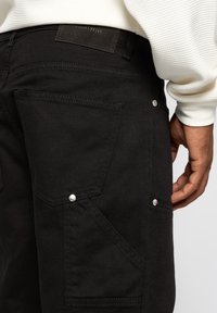 Black denim cargo pants featuring multiple pockets, silver hardware accents, and a textured fabric. The waistband includes a label visible at the back.