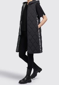 Black quilted sleeveless vest with a hood, side pockets, and contrasting interior. Paired with black top and ankle boots.