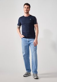 Camiseta azul marino con cuello redondo y un pequeño logo; presenta detalles a rayas rojas y blancas en las mangas. Combinada con vaqueros claros y zapatillas.