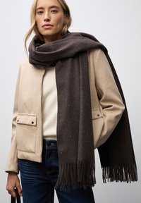 Brun uldhalstørklæde med frynser, kombineret med en beige jakke og en cremet sweater, der skaber et lagdelt look passende til koldere vejr.