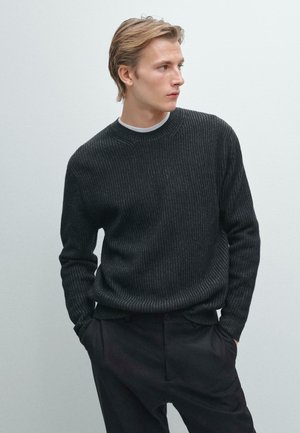 Maglione a coste scuro con collo a giro, realizzato in materiale a maglia texturizzato. Abbinato a pantaloni scuri, il modello si erge su uno sfondo neutro.