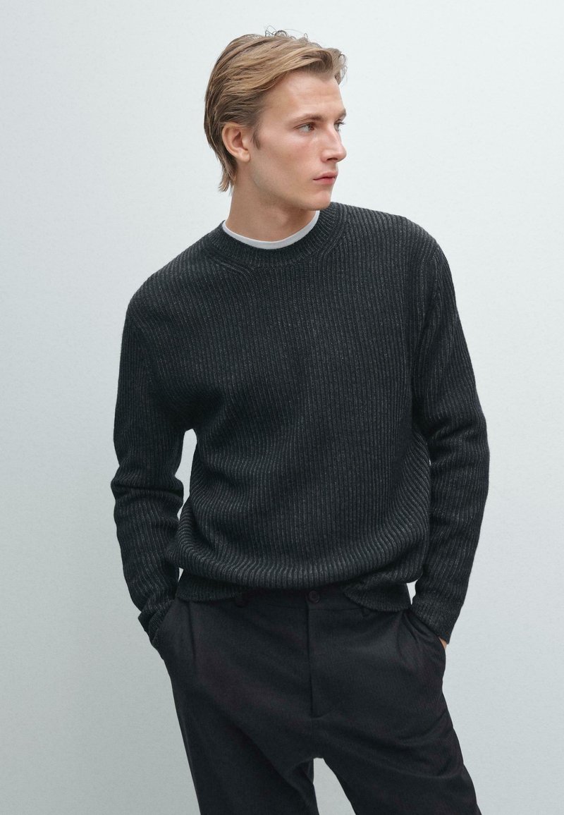 Maglione a coste scuro con collo a giro, realizzato in materiale a maglia texturizzato. Abbinato a pantaloni scuri, il modello si erge su uno sfondo neutro.