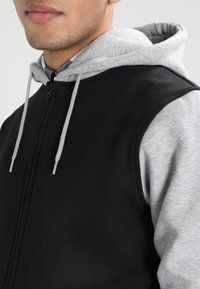 Gilet nero con cappuccio grigio sotto. Presenta un tessuto bicolore, una texture liscia e dettagli con lacci.