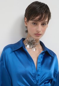 Una persona con i capelli corti e scuri indossa una camicia di raso blu lucido e un grande collare floreale in argento su uno sfondo chiaro e semplice.