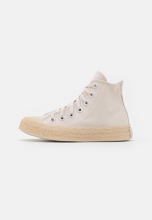 Beige hoge canvas sneaker met een getextureerde rubberen zool, metalen oogjes en beige veters, zijaanzicht tegen een witte achtergrond.