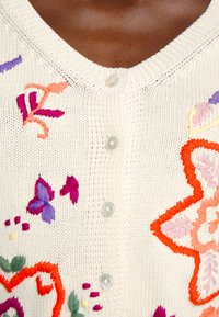 Strickjacke in Creme mit bunten gestickten Blumen und Schmetterlingen; verfügt über einen V-Ausschnitt und fünf klare Knöpfe vorne.