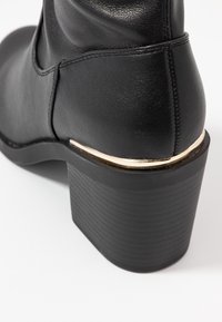 Botín de cuero negro hasta el tobillo con un tacón cuadrado y un delgado detalle dorado encima de la suela, mostrado desde detrás sobre un fondo blanco.