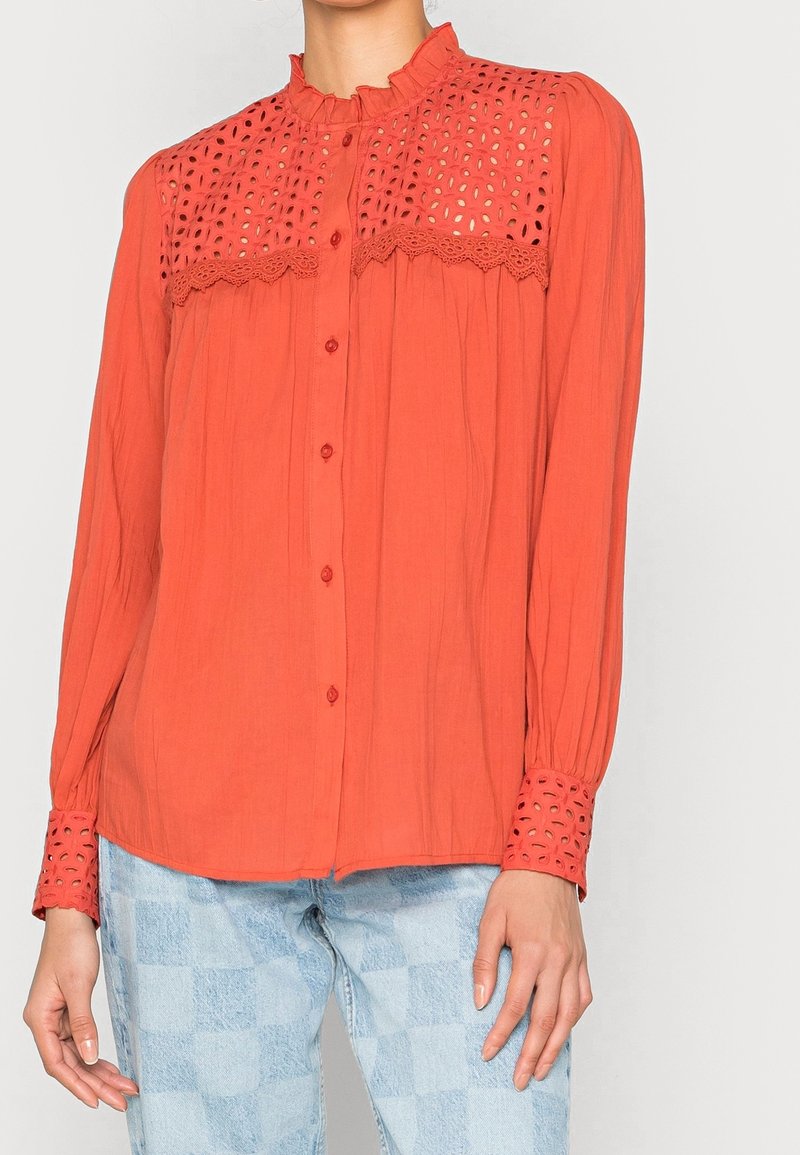 Blouse orange à manches longues avec un col à volants, des détails en broderie anglaise sur le corsage et des poignets à motif ; tissu doux avec une coupe décontractée.