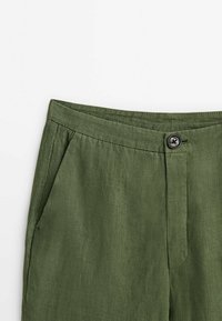 Pantalon en lin vert olive avec poches latérales, fermeture par bouton et détail de couture à la taille sur fond blanc.