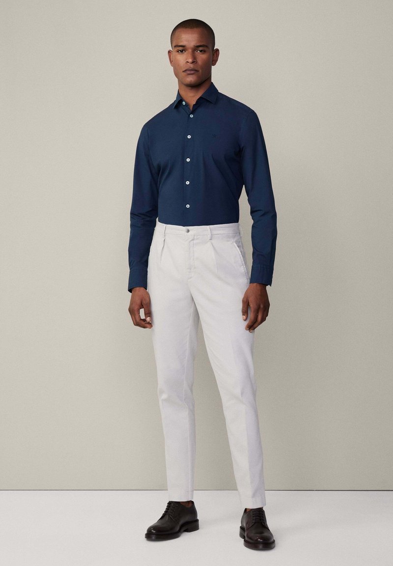 Marineblaues Langarmshirt mit Knöpfen und spitzem Kragen, kombiniert mit weißen Hosen und schwarzen Schuhen. Glatte Stoffqualität, schmaler Schnitt.