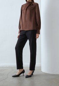 Blusa marrone a maniche lunghe con colletto alto e dettagli plissettati sul davanti; abbinata a pantaloni neri su misura e tacchi a punta neri.
