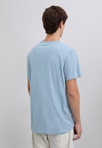 Hombre con cabello corto castaño que lleva una camiseta de manga corta azul claro y pantalones blancos, de espaldas frente a un fondo liso.