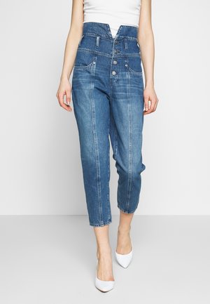 Femme portant un jean en denim bleu taille haute avec des boutons à l’avant, des coutures verticales et des talons blancs à bout pointu, debout devant un fond uni.