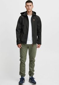 Sort softshell-jakke med hætte, lynlås foran og logo på brystet; parret med olivengrønne bukser og marineblå træningssko. Enkelt design.