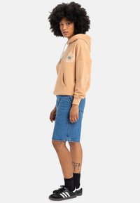 Beige Kapuzenpullover mit Grafikdruck, Fronttasche und Kordelzug-Details, kombiniert mit einem blauen Denim-Bleistiftrock und schwarzen Adidas Sneakern.