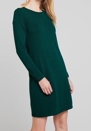 Femme portant une robe longue à manches longues, de couleur vert foncé, longueur genou, debout devant un fond gris clair uni.
