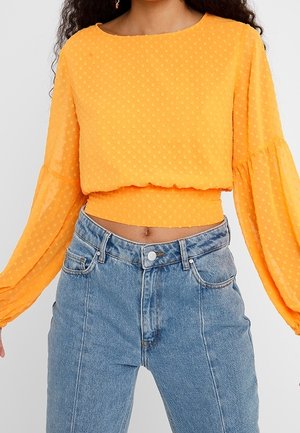 Frau trägt ein leuchtend oranges langärmeliges Crop-Top mit strukturierten Punkten und hoch taillierte hellblaue Jeans.