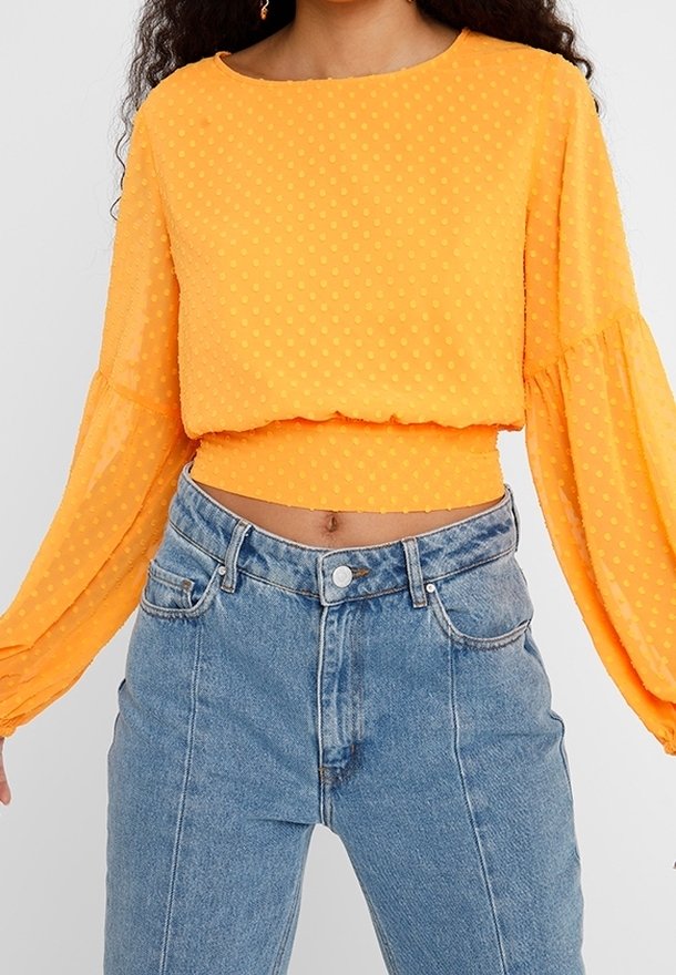 Femme portant un crop top orange vif à manches longues avec des points texturés et un jean taille haute bleu clair.