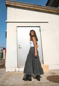 Ilgas pilkas sluoksniuotas sijonas, pagamintas iš džinsinio audinio, derinamas su baltu marškinėliais be rankovių ir juodais auliniais batu, prieš paprastą sieną ir aiškiai mėlyną dangų.